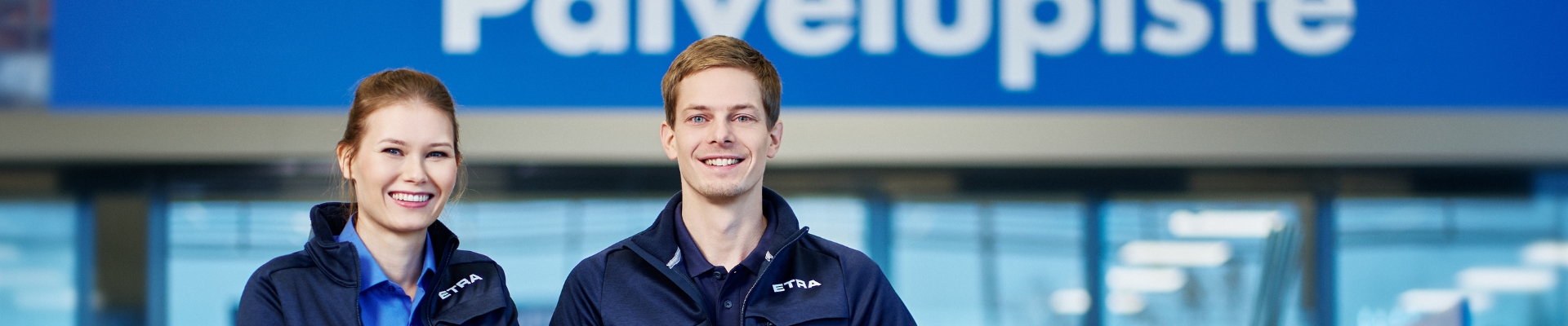 Two smiling ETRA employees in uniform standing in front of the customer service point, marked with a large 'Palvelupiste' sign in Finnish. The background shows a modern retail or service environment.