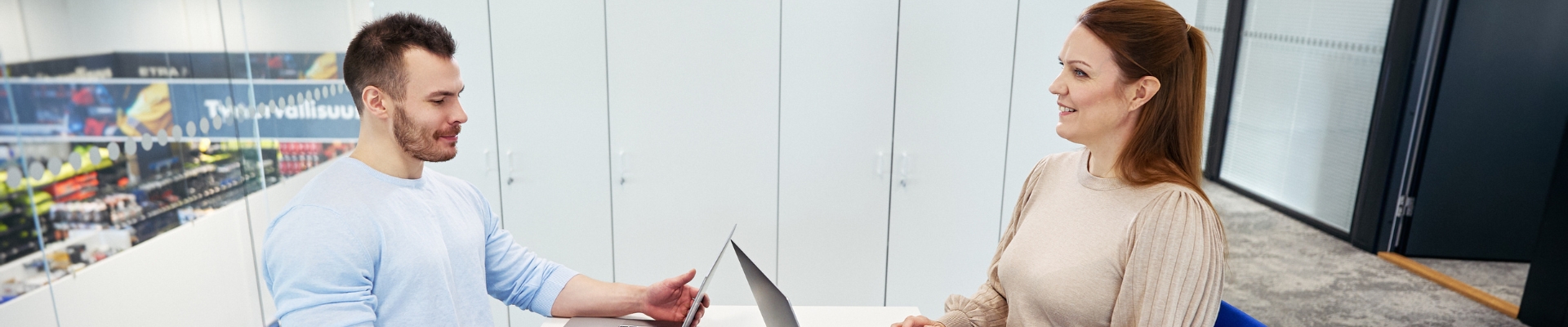 Two colleagues sitting across from each other at a table in a modern office meeting room, both working on laptops and engaged in conversation.