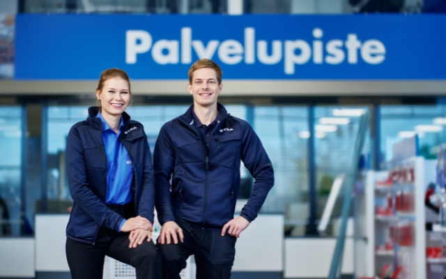 Two smiling ETRA employees in uniform standing in front of the customer service point, marked with a large 'Palvelupiste' sign in Finnish. The background shows a modern retail or service environment.