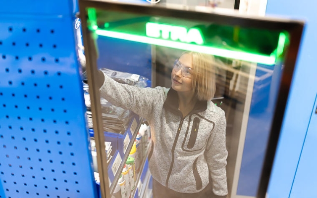 A woman wearing safety glasses reaches for a product inside a blue ETRA vending or storage cabinet, viewed through a transparent door with a glowing green ETRA logo.