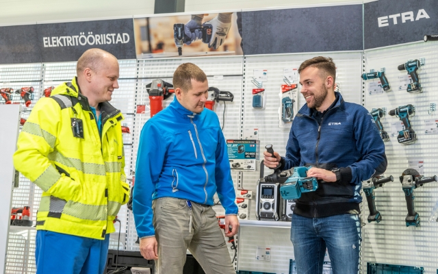 Three men standing in front of a power tools display at an Etra store. One of them, wearing an Etra-branded jacket, is demonstrating a power tool to the others.