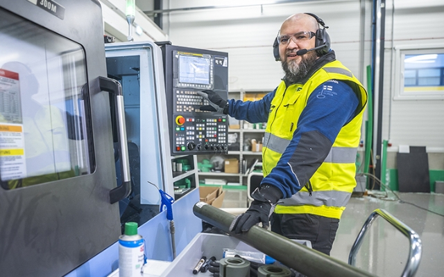 A worker wearing a high-visibility Etra vest and protective headset operating a CNC machine in an industrial workshop. He is smiling while using the machine's control panel, with tools and equipment visible around him.