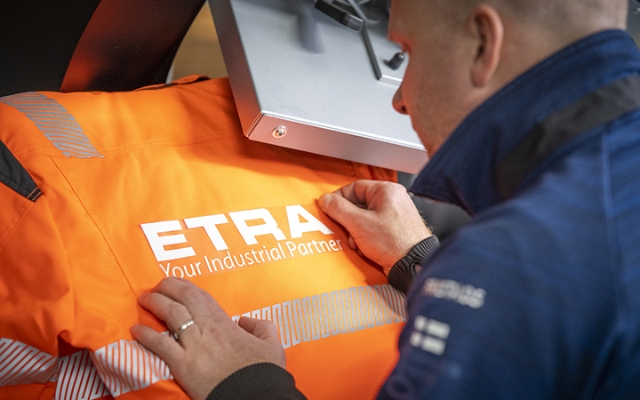 A person carefully aligning an orange high-visibility work jacket under a machine, featuring the ETRA logo and the slogan 'Your Industrial Partner' being applied to the fabric.