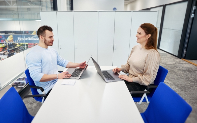 Two colleagues sitting across from each other at a table in a modern office meeting room, both working on laptops and engaged in conversation.