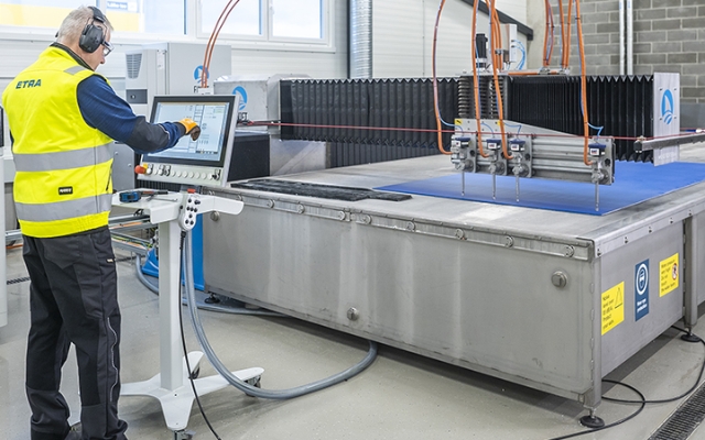 An Etra employee operating a CNC waterjet cutting machine via a touchscreen control panel, wearing safety gear including ear protection and a high-visibility vest, in an industrial manufacturing environment.