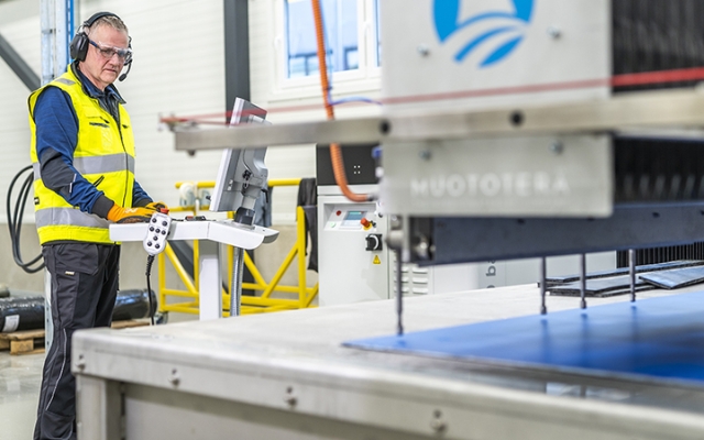 An Etra employee wearing safety glasses, hearing protection, gloves, and a high-visibility vest operates a CNC waterjet cutting machine from a control panel in an industrial workshop.