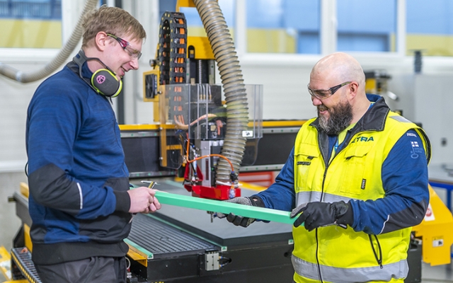 Kaks töötajat CNC-seadme juures vahetamas plastdetaili, kandes tööriideid ja kaitsevahendeid.