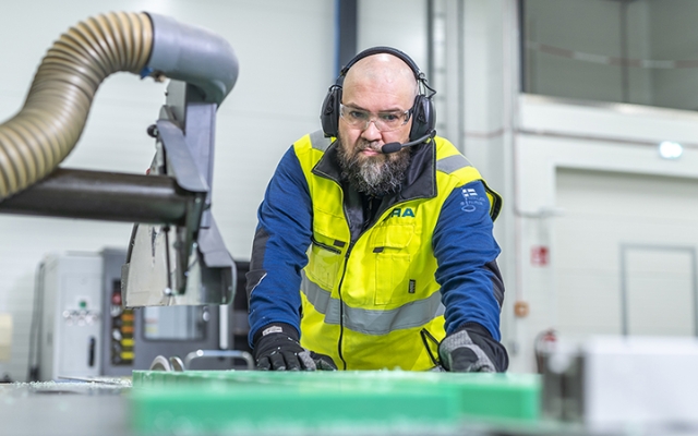 Töötaja kõrvaklappide ja mikrofoniga töötlemas rohelist plastplaati tööstusliku saega.