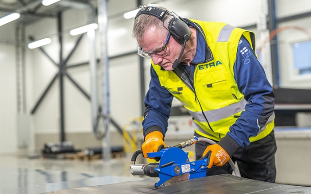 Etra employee using a handheld tool to finish the edges of a plastic sheet, wearing safety glasses, earmuffs, and a high-visibility vest.