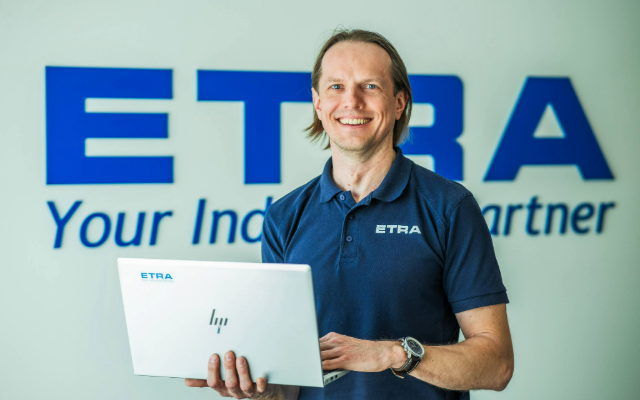 Etra töötaja naeratab ja hoiab käes sülearvutit, taustal ettevõtte logo ja loosung “Your Industrial Partner”.
