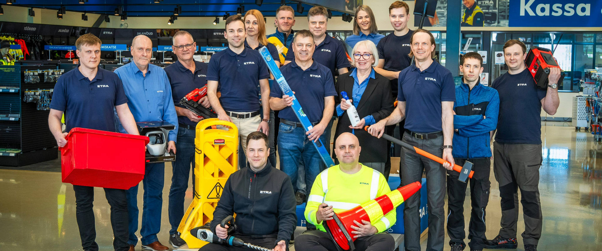 The Etra team at the store’s occupational safety section. Employees pose with various industrial products, such as warning cones, containers, tools, and safety equipment. In the background, signs read “Occupational Safety” and “Checkout”.