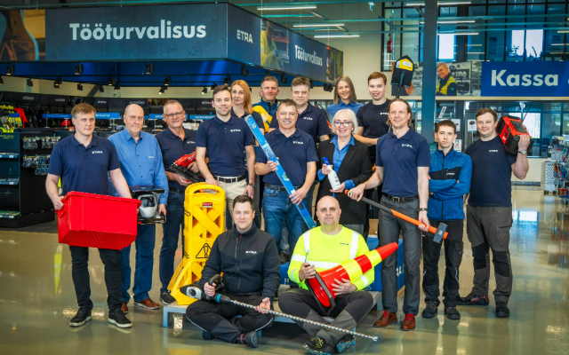 The Etra team at the store’s occupational safety section. Employees pose with various industrial products, such as warning cones, containers, tools, and safety equipment. In the background, signs read “Occupational Safety” and “Checkout”.